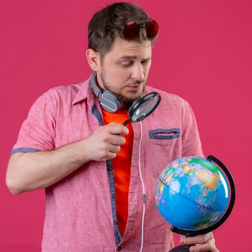 young-traveler-man-with-headphones-looking-globe-through-magnifying-glass-standing-pink-background_141793-20393